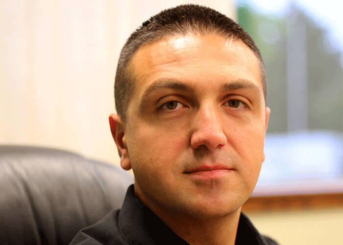 A man with short dark hair sitting in an office chair, wearing a black collared shirt, looking at the camera with a neutral expression.
